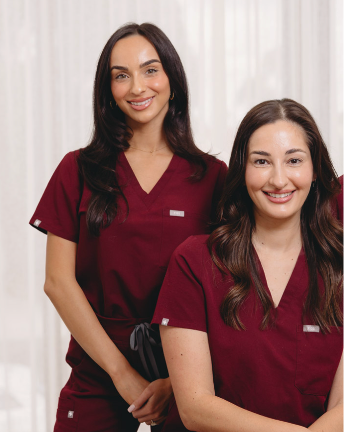 Two women wearing matching burgundy V-neck scrub tops and pants in a bright indoor setting with white curtains in the background.