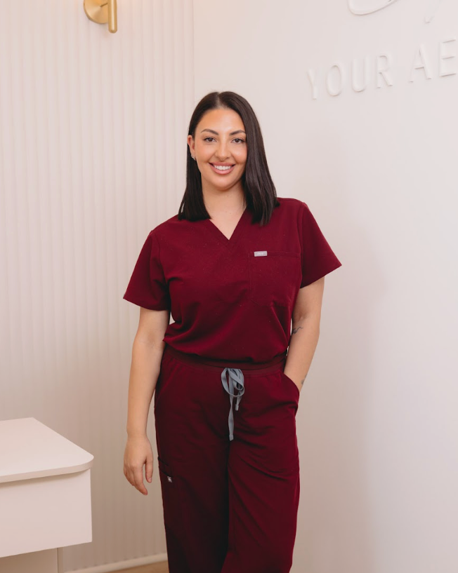 A woman wearing a matching burgundy scrub top and scrub pants set with a gray drawstring, standing in a brightly lit clinical-style room with a white wall and wood floor.