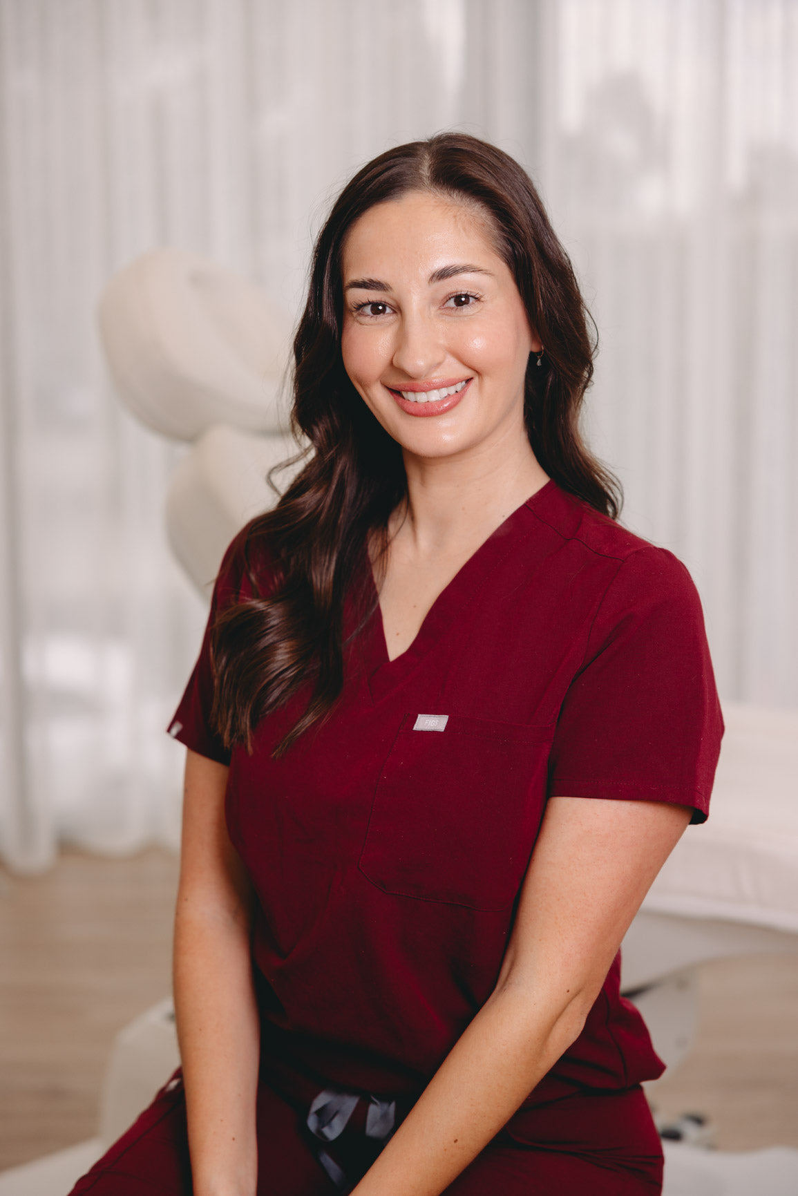 A smiling woman with long dark brown hair wearing burgundy medical scrubs, seated in a brightly lit, modern room with white curtains and a treatment bed in the background.