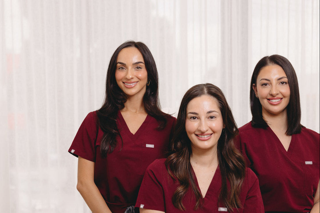 Three women wearing matching burgundy medical scrubs uniforms, with one seated in the center and two standing behind her, in a bright, modern white clinical room.