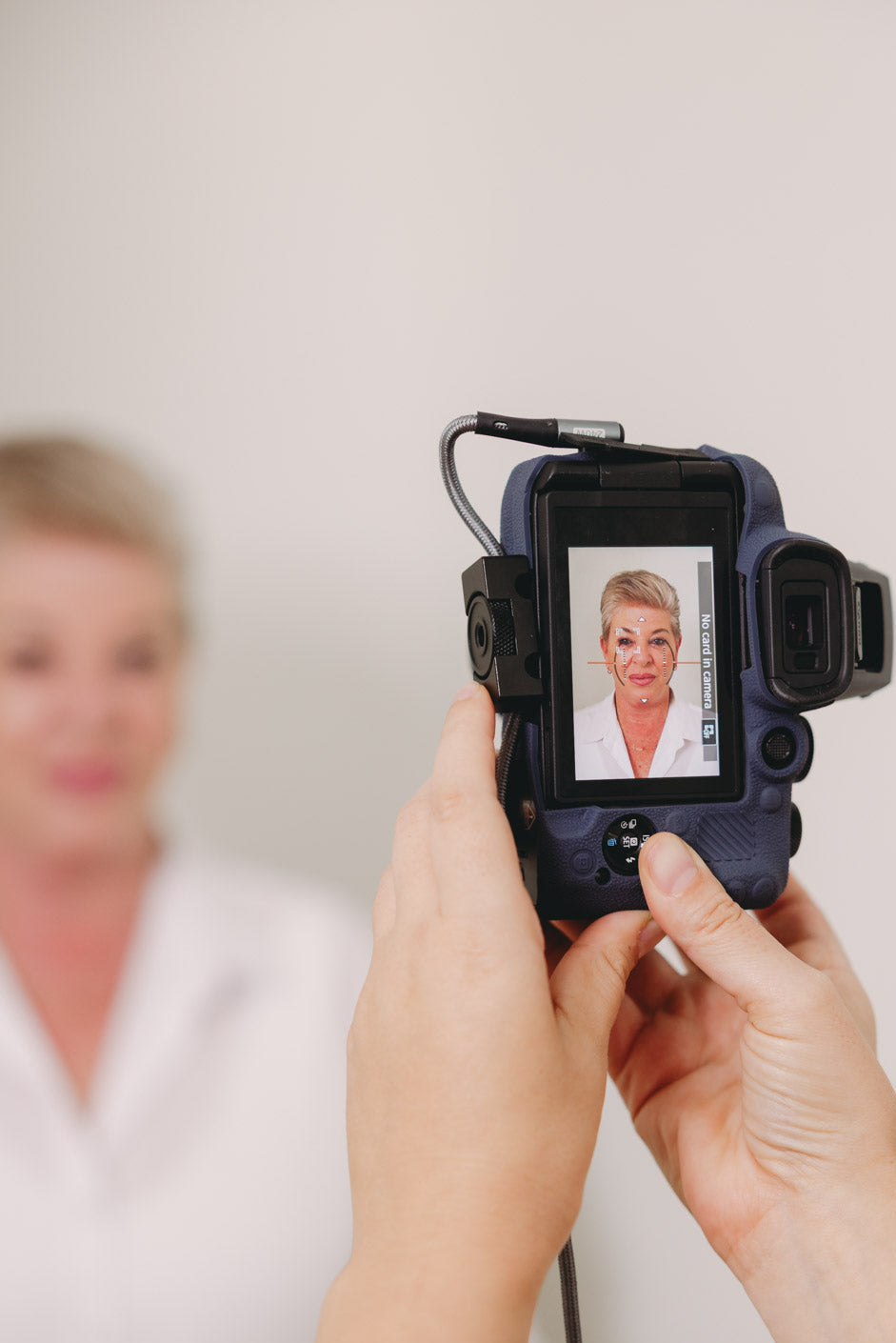 A close-up of a person holding a black digital camera with a screen showing a portrait of an older woman, with another woman blurred in the background.